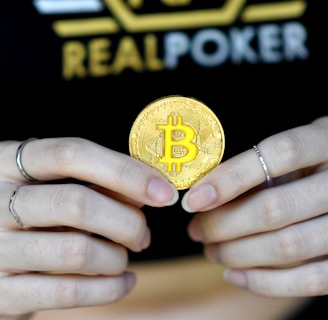 A person holds a golden coin with a Bitcoin symbol between their fingers. The background features the word 'REALPOKER' in bold letters on a dark shirt.