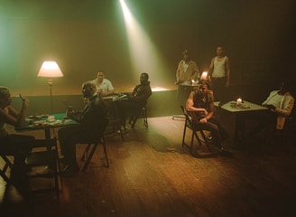 Several people sit at tables in a dimly lit room with a warm, moody atmosphere. A lamp and candle provide soft lighting, casting shadows across their faces. Cards and chips are on the tables, suggesting a game of poker. The setting is rustic, with wooden floors and simple furniture.